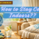 Landscape image of a summer living room with a blue fan, glass of ice water, and sunlight streaming through sheer curtains. Text overlay reads “How to Stay Cool Indoors??”
